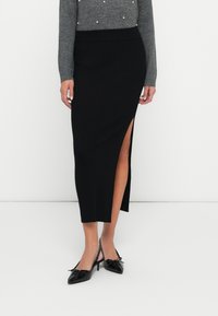 Falda midi negra acanalada con una abertura alta en un lado, combinada con zapatos negros de punta. El material parece suave con un diseño ajustado.
