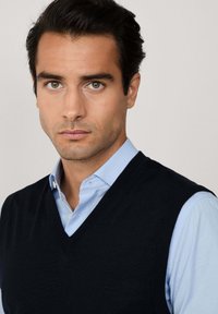 Muž s tmavými vlasmi, oblečený v svetlo modrej košeli s golierom a čiernym bezrukávovým sveterom s výstrihom do V, čelí dopredu s neutrálnym výrazom.