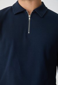 Pullover azul-marinho com colarinho com fecho éclair, feito de tecido canelado, com um fecho de metal prateado e uma superfície texturizada.