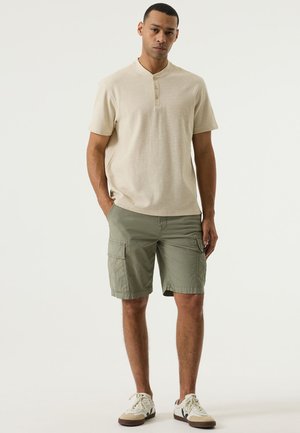 Beige henley-shirt met korte mouwen en een drieknops sluiting, gecombineerd met olijfgroene cargoshorts tot halverwege de dij met zakken aan de zijkant. Witte en beige sneakers.