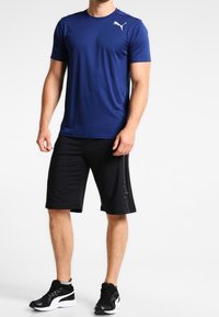 Modrá sportovní košile s krátkým rukávem a bílým logem na levé straně hrudi, v kombinaci s černými kraťasy po kolena a černými sportovními botami.