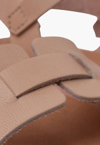 Beige ādas čības ar platām, plakaniem siksnām un gludu tekstūru. Siksnu malās redzama šuvju detaļa.