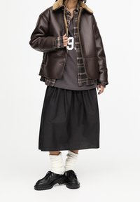 Veste en cuir marron avec col en shearling, chemise à carreaux en dessous, tee-shirt gris, jupe noire et chaussures à lacets noires avec des chaussettes blanches texturées.