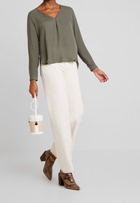 Olivegrünes, langärmliges Blusenshirt mit V-Ausschnitt und einem subtilen Faltenwurf, kombiniert mit cremefarbenen, locker sitzenden Hosen. Braune Ankle-Boots im Schlangendruck.