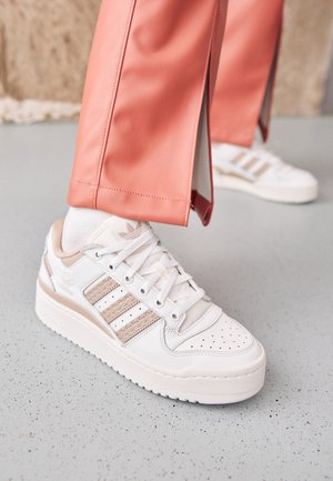 Osoba nosząca białe i beżowe sneakersy Adidas z białymi skarpetkami oraz koralowymi spodniami z bocznymi rozcięciami stoi na szarej, cętkowanej podłodze.