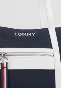 Marinblå tote-väska med vita läderdetaljer, silverfärgad dragkedja och "TOMMY"-logotyp framtill. Har en randig nyckelringsdetalj.