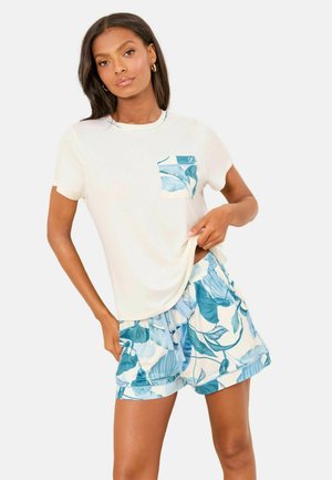 Moterys baltais trumpomis rankovėmis marškinėliais su mėlynos spalvos kišene su lapų raštu ir atitinkančiais mėlynos ir baltos spalvos lapų rašto šortais, stovinti priešais baltą foną.