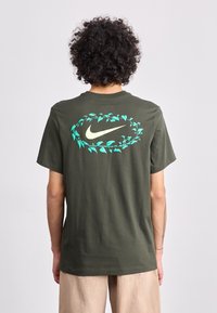 Oliven grøn bomuld t-shirt med et stort, stiliseret Nike-logo omgivet af teal blade på ryggen. Korte ærmer, afslappet pasform.