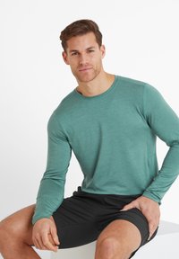 Man with short hair wearing a teal long-sleeve shirt and black shorts, sitting on a white block against a white background.