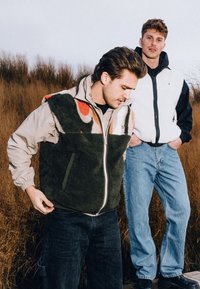 Dva modely majú na sebe vesty; jeden v zelenej flísovej veste s farebným vzorovaným horným dielom a druhý v čierno-bielej flísovej veste. Obe modely majú na sebe džínsy.