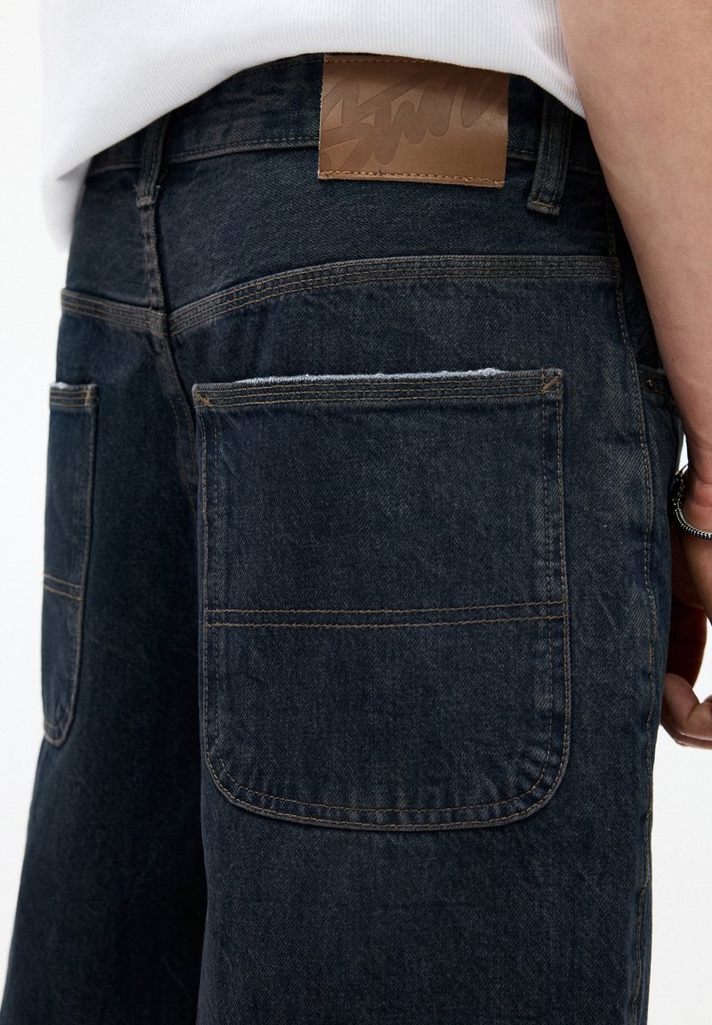Gros plan sur un short en denim foncé avec de grandes poches arrière, une étiquette de marque en cuir marron sur la ceinture, porté par une personne vêtue d'une chemise blanche.