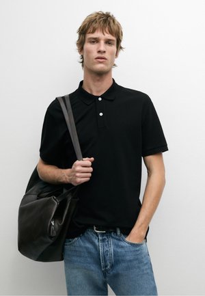 Jonge man in een zwart poloshirt en blauwe spijkerbroek houdt een zwarte leren tas over zijn schouder tegen een effen witte achtergrond.