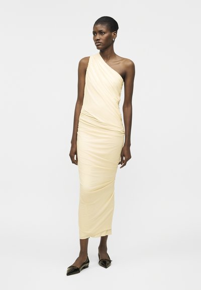 Sieviete stāv, tērpusies vienas pleca dzeltenā svārku vidēja garuma kleitā ar rušām, valkā melnas smailas dzīvokļu kurpes, fonā redzams gaišs vienkāršs fons.