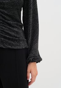 Top nero con un design texturizzato caratterizzato da fili metallici, maniche lunghe con polsini arricciati e vestibilità rilassata. Pantaloni neri parzialmente visibili.