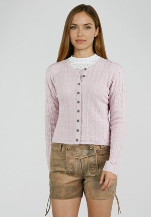 Vrouw die een lichtroze gebreid vest met knopen draagt over een witte kanten top, gecombineerd met traditionele bruine leren lederhosenshorts met geborduurde details.