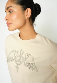 Felpa beige con collo a girocollo a costine. Presenta un design di ali e simboli in strass metallici sulla parte frontale. Tessuto morbido al tatto.