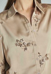 Camisetta in satin beige con colletto, decorata con applicazioni floreali di paillettes in bronzo e marrone chiaro, e chiusura frontale con bottoni.