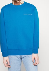 Sudadera azul con cuello redondo, puños y dobladillo acanalados, presenta un logo bordado en gris. Combinada con jeans de mezclilla azul claro.