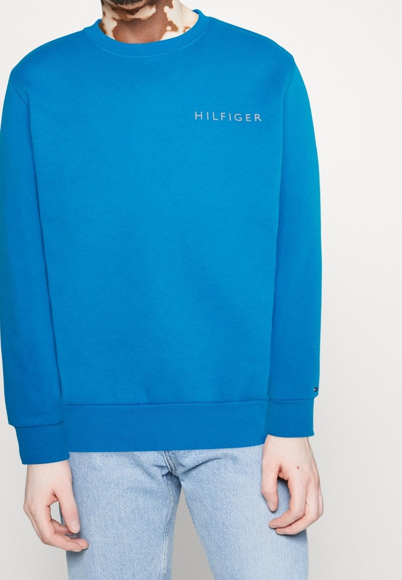 Sudadera azul con cuello redondo, puños y dobladillo acanalados, presenta un logo bordado en gris. Combinada con jeans de mezclilla azul claro.