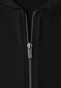 Musta vetoketjullinen takin yksityiskohta, jossa kiiltävä metallivetoketju ja teksturoitu kaulus, sileän kangaspohjan edessä.