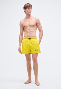 Dzelteni peldēšanas šorti ar aukliņu jostasvietā, sānos redzama kontrastējoša melna logo. Gluda tekstūra, ceļam garš dizains.