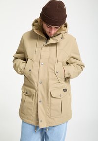 Beige insulated parka jacket with buttoned front, drawstring hood, and multiple front pockets; lined with soft textured material.