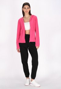 Felpa rosa con zip e cappuccio, abbinata a pantaloni jogger neri e sneakers bianche. Il tessuto sembra morbido e ha una vestibilità comoda.