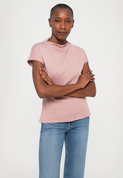 Pink short-sleeve top with a high neckline, smooth texture, and fitted design. Paired with light blue denim jeans.