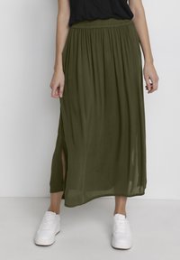 Jupe maxi vert olive en tissu léger et fluide avec une ceinture élastique et une fente latérale, portée avec des baskets blanches.