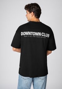 Schwarzes übergroßes T-Shirt mit weißem Text "DOWNTOWN CLUB" und "RISK IT ALL FOR YOUR PASSION" auf der Rückseite. Kurze Ärmel, aus Baumwolle.