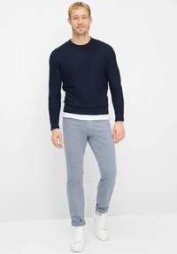Glimlachende man die een marineblauwe trui draagt over een wit overhemd, lichtgrijze broek met omslagen en witte sneakers, staand tegen een witte achtergrond.