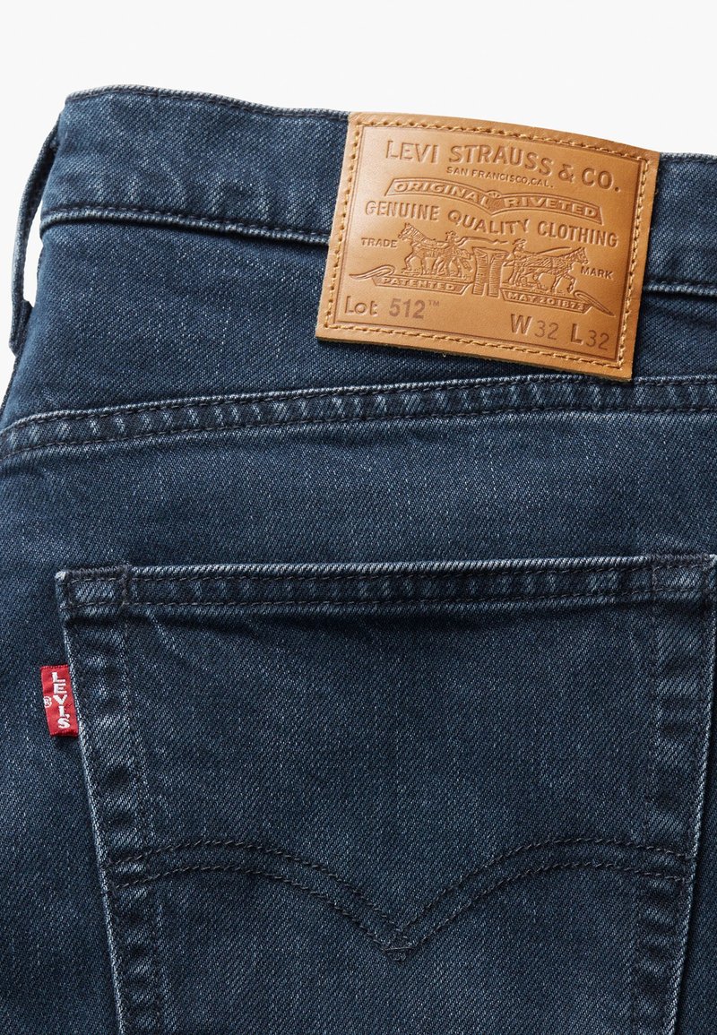 Dunkelblaue Denim-Jeans mit einem Lederaufnäher auf der Rückseite, der eingestanzten Text trägt. Auffälliges rotes Levi's-Label an der Seite. Klassisches Fünf-Taschen-Design.