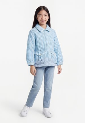 Pige stående og smilende, iført lyseblå hundetandsjakke, lyseblå jeans og hvide sneakers mod en ensfarvet baggrund.