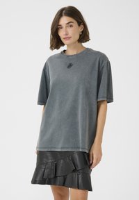 Tricou din bumbac gri supradimensionat, cu guler rotund și mâneci scurte, având un mic design brodat negru pe piept, asortat cu o fustă din piele neagră cu volane.