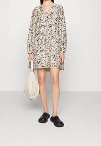Robe à motifs floraux, longueur genou en tissu léger avec un décolleté en V et des manches longues. Associée à des sabots noirs et un sac en tissu beige.
