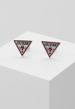 Paire de boucles d'oreilles à clous triangulaires de la marque Guess en blanc et rouge avec un point d'interrogation et le texte "WASHED JEANS" sur une surface blanche.