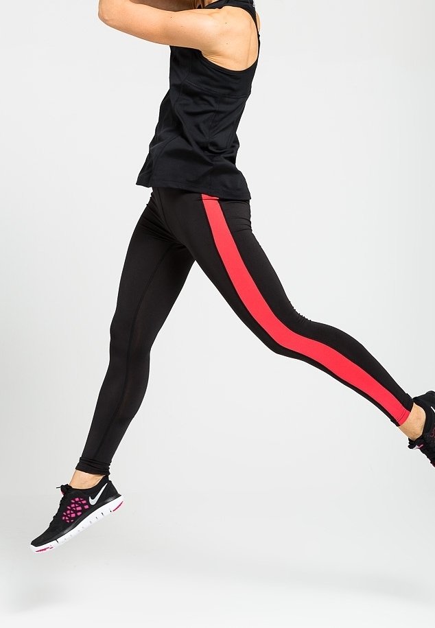 Personne portant un legging noir avec une bande rouge et des chaussures de sport noires, capturée en plein saut sur un fond clair uni.