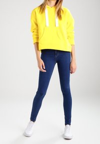 Felpa gialla con cappuccio e cordini bianchi, tasca frontale, abbinata a jeans skinny blu scuro e sneakers bianche. Texture del tessuto liscia.