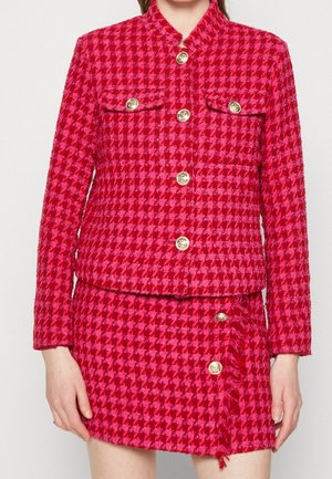 Kobieta ubrana w czerwoną i różową kurtkę w kratę węgierską oraz pasującą spódnicę z złotymi guzikami, stojąca na jednolitym tle.