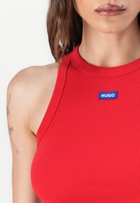 Débardeur rouge côtelé avec un col haut et une étiquette bleue "HUGO". Le tissu est lisse avec une coupe ajustée et une texture subtile.