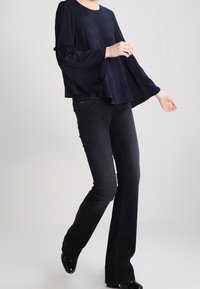 Top navy a maniche a campana in tessuto testurizzato, abbinato a jeans neri a zampa d'elefante. Le scarpe hanno una finitura lucida e l'outfit presenta dettagli di design discreti.