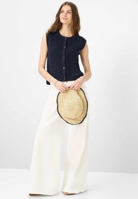 Donna che indossa un gilet senza maniche blu navy con bottoni e pantaloni larghi bianchi, che tiene in mano un cappello di paglia intrecciata con bordatura nera.