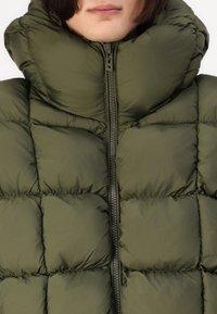 Jachetă puffer de culoare verde măslin cu secțiuni mari matlasate, guler înalt și fermoar frontal. Țesătură de nailon netedă cu un finisaj mat.