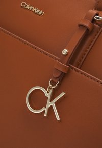 Brun faux läderhandväska med en guldig "CK" charm som är fäst med ett band. Har präglad logotyp och stickade detaljer.