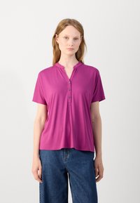 Fuksia lyhythihainen henley-paita, jossa on kaulus, kolme napin sijaa ja pehmeä, sileä kangas. Yhdistetty löysiin sinisiin farkkuihin.