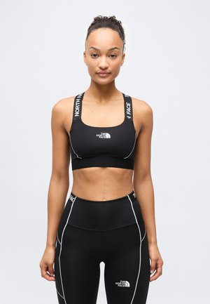 Kvinne vendt fremover iført svart sports-bh og matchende leggings fra The North Face med hvit logo og sømmer på en ensfarget lys grå bakgrunn.