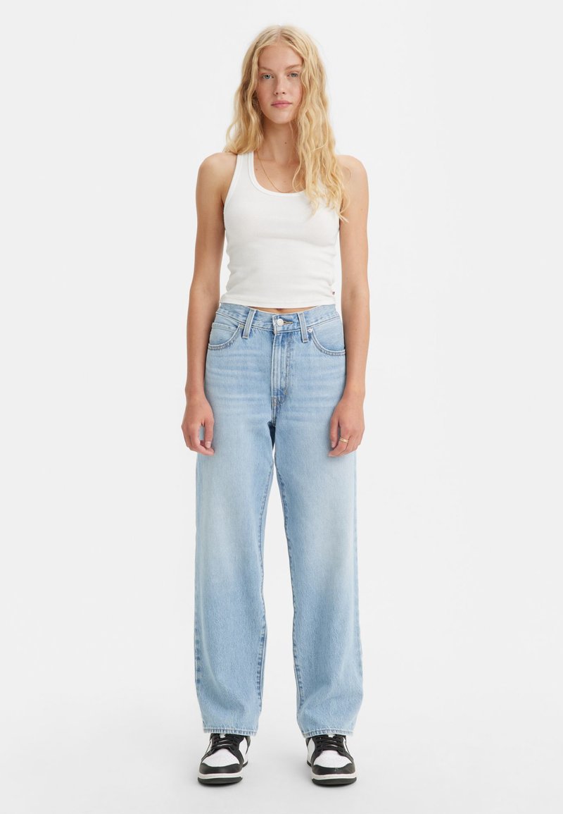 Helle blaue Jeans mit hoher Taille, geraden Beinen und dezentem Verblassen. Kombiniert mit einem weißen Tanktop und schwarz-weißen Turnschuhen.