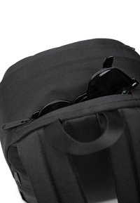 Mochila negra con un exterior de tela texturizada, asa superior y gafas de sol visibles en un bolsillo con cremallera. Diseño minimalista.