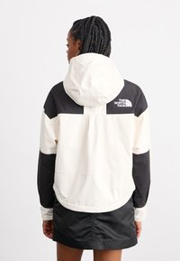 Kvinne med flettet hår iført en svart og hvit hettejakke fra North Face som vender bort fra en ensfarget hvit bakgrunn.