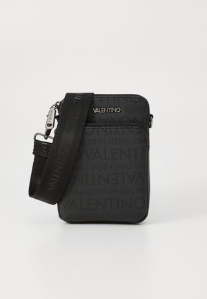 Bolso cruzado de tela negra con superficie texturizada, que presenta un logotipo en relieve y una correa desmontable. Cierre con cremallera y diseño minimalista.